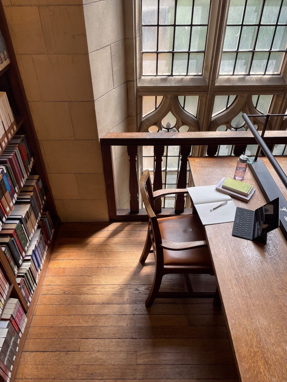 An old wooden desk in the Wills Memorial Library, next to a gothic style window. On the desk are a couple of books, notebook and tablet computer.